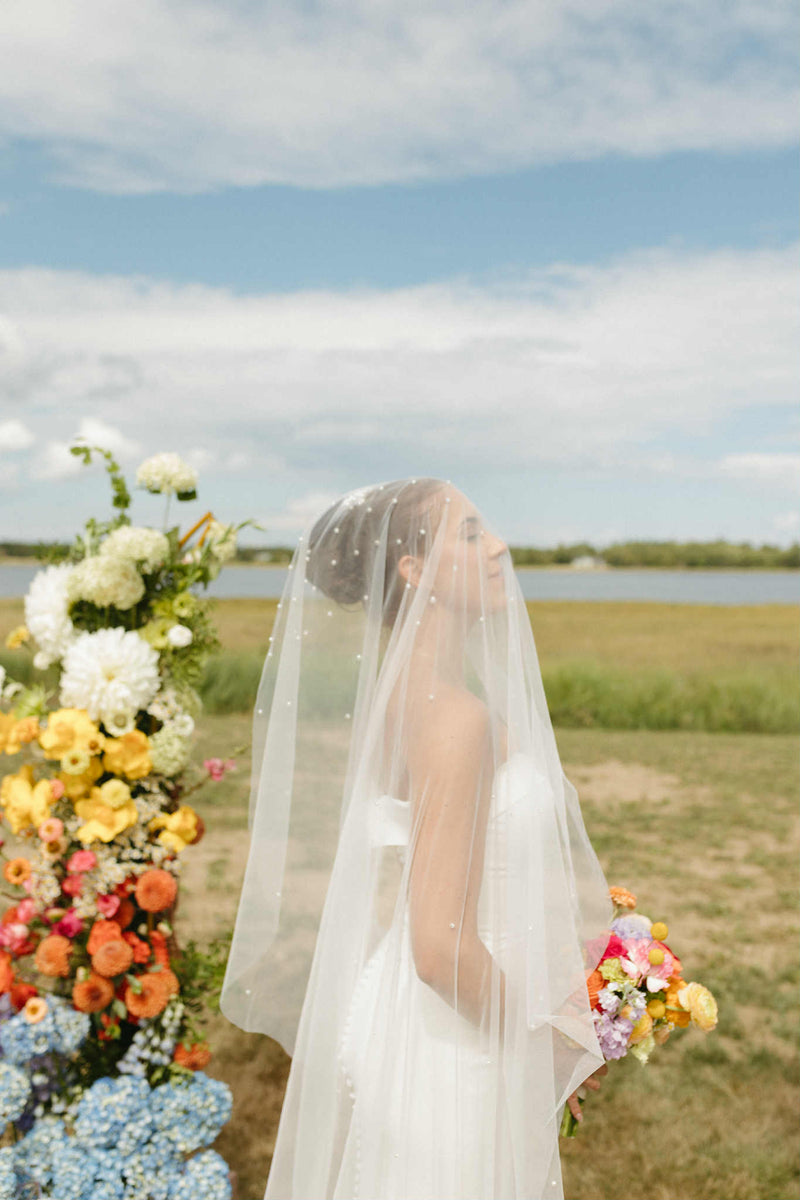 Bride in a white dress with a floral veil standing in a field with wedding flowers and a lake in the background. Flowers by Primavera Floral studio a wedding florist in Moncton new brunswick.