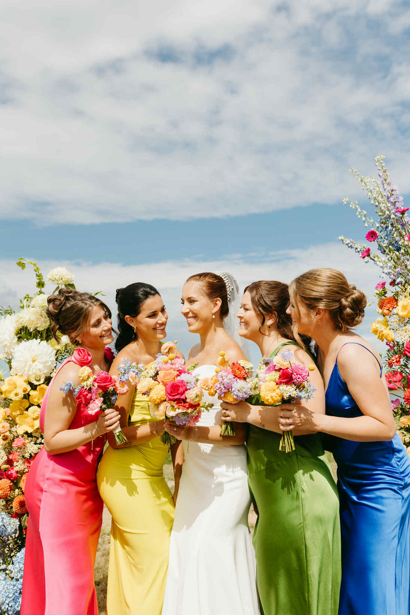 Bride with four bridesmaids in colorful dresses holding flowers bouquet in Moncton against a scenic flower background made by Primavera floral studio in Moncton new brunswick serving wedding flowers in Moncton and beyond.