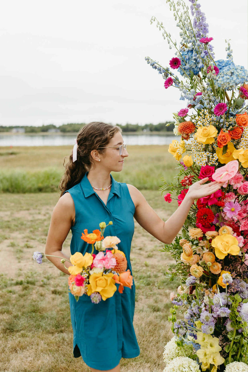 Julie Losier from Primavera floral studio, wedding florist, in a blue dress interacting with a vibrant floral arrangement for weddings outdoors in Moncton.